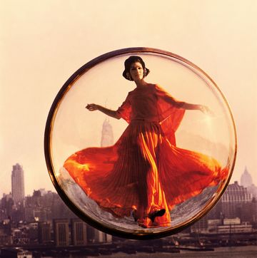 a woman in an orange dress poses in a bubble floating above new york